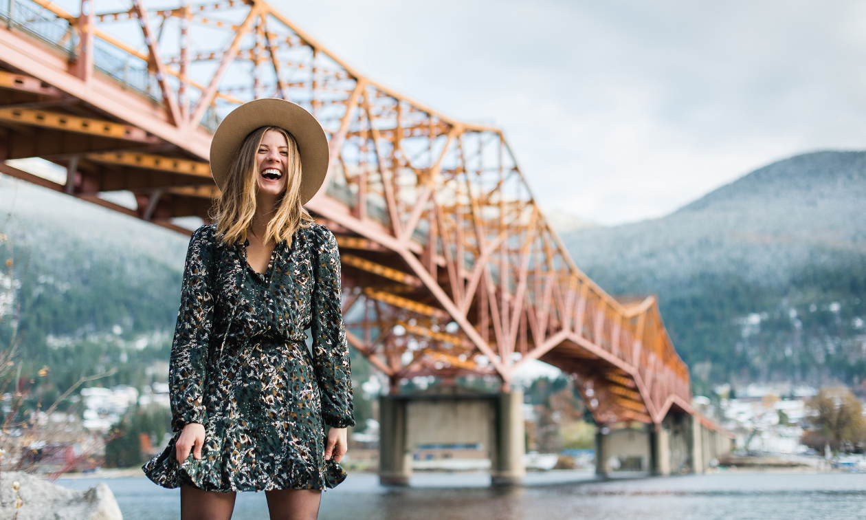 Amanda Mary laughs while wearing a dress in front of an orange bridge in Nelson, B.C.