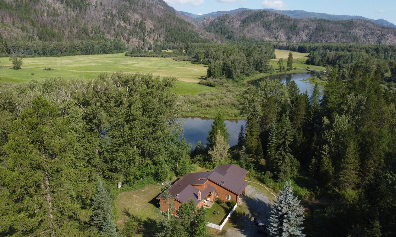 An aerial shot of a large brown Kootenay home in the midst of trees, mountains and rivers. 