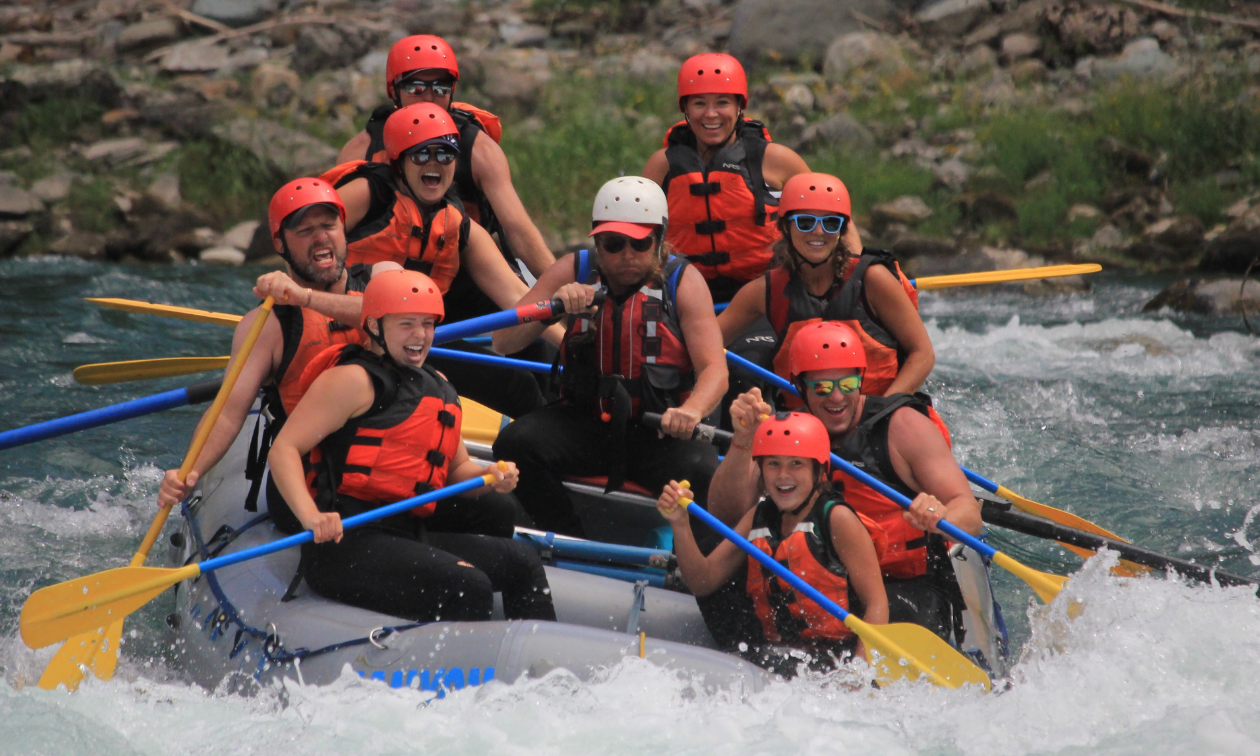 A group of white water rafters smile as they dip into deep water on a raft. 