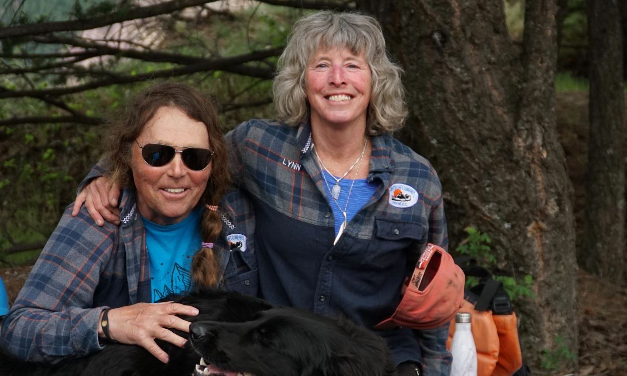 Blair Craig has long brown hair and large sunglasses. He has his arm around Lynn Muller, who has grey hair and smiles with a dark blue shirt on. 