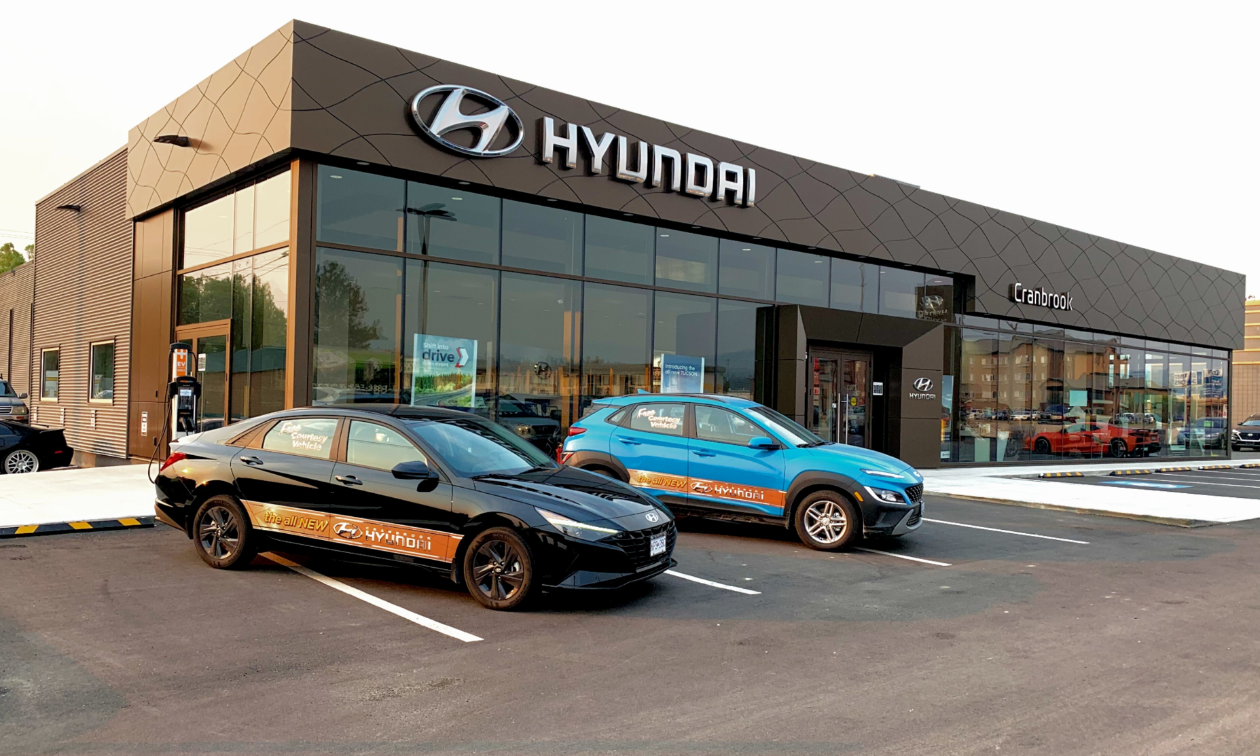 Two cars, one black, one blue, are parked in front of Cranbrook Hyundai’s new showroom with large glass windows. 