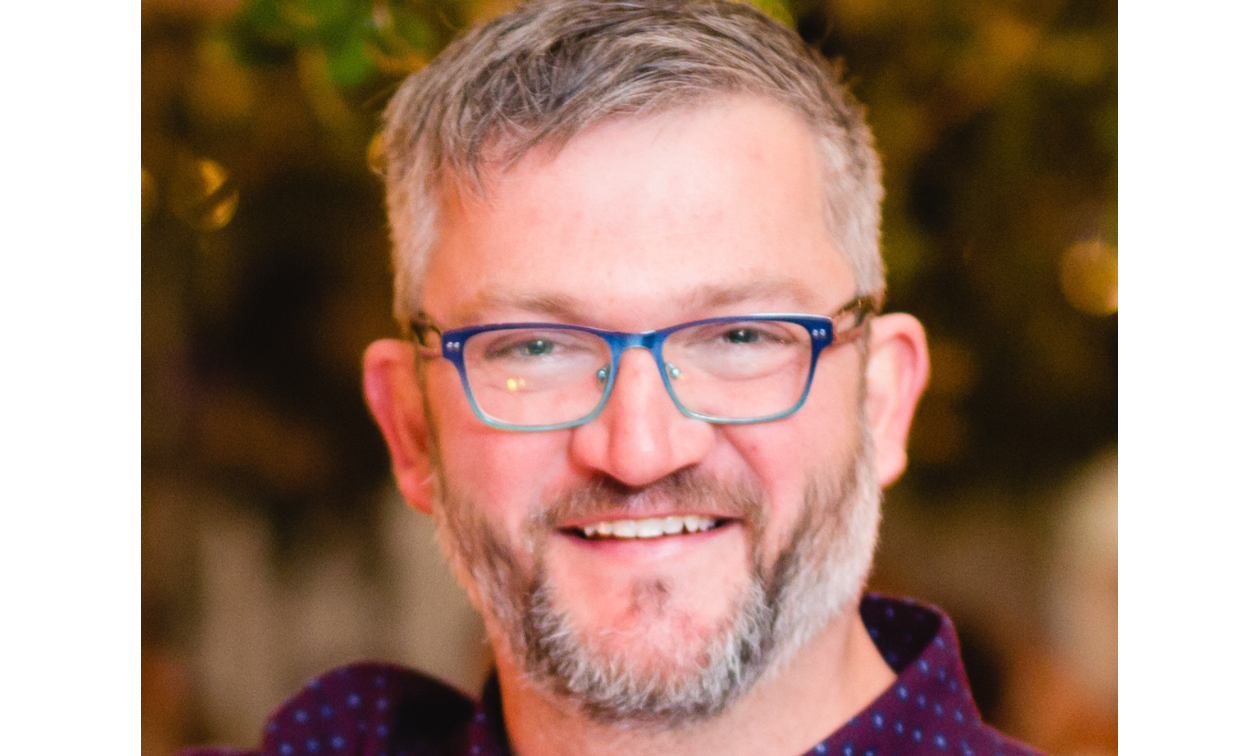 Daniel Holden smiles, wearing black-rimmed glasses and a salt and pepper beard. 