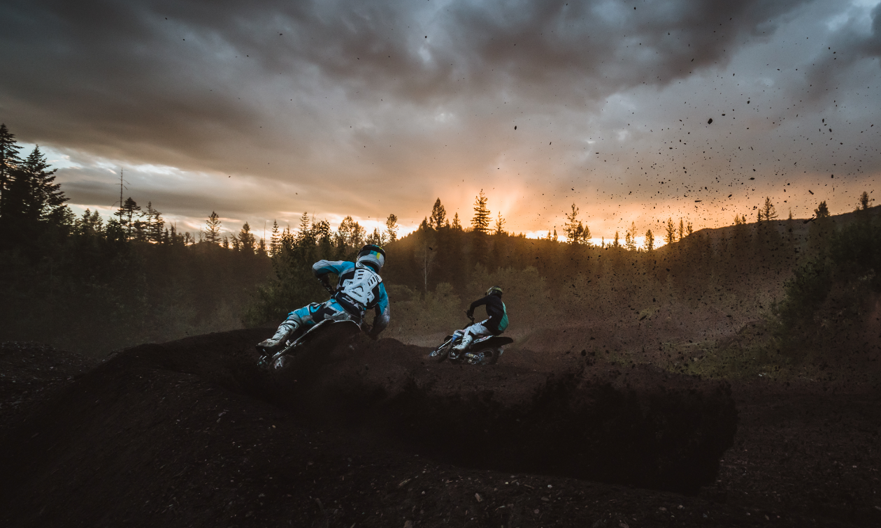 Two dirt bikers take a sharp turn at the West Kootenay Recreational Dirt Bike and ATV Society motocross track at dusk.