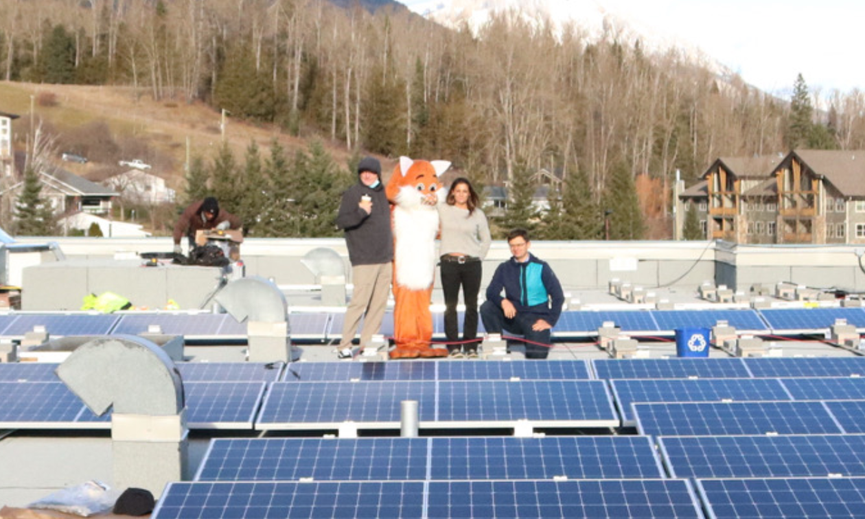 Four people stand on a roof covered in solar panels. One of the people is dressed up like an orange fox mascot. 