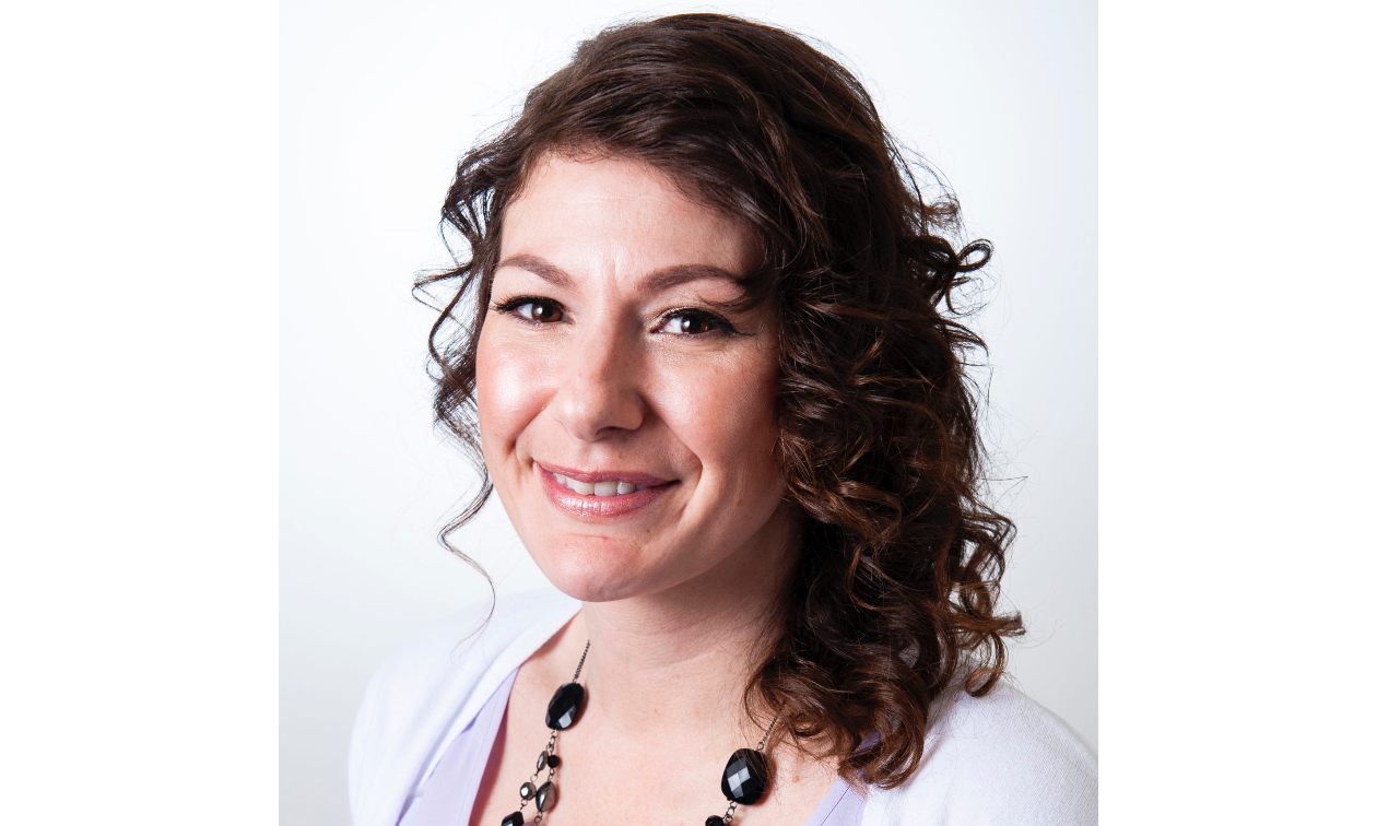 Helen Barron smiles. She has long curly brown hair and wears a white shirt. 