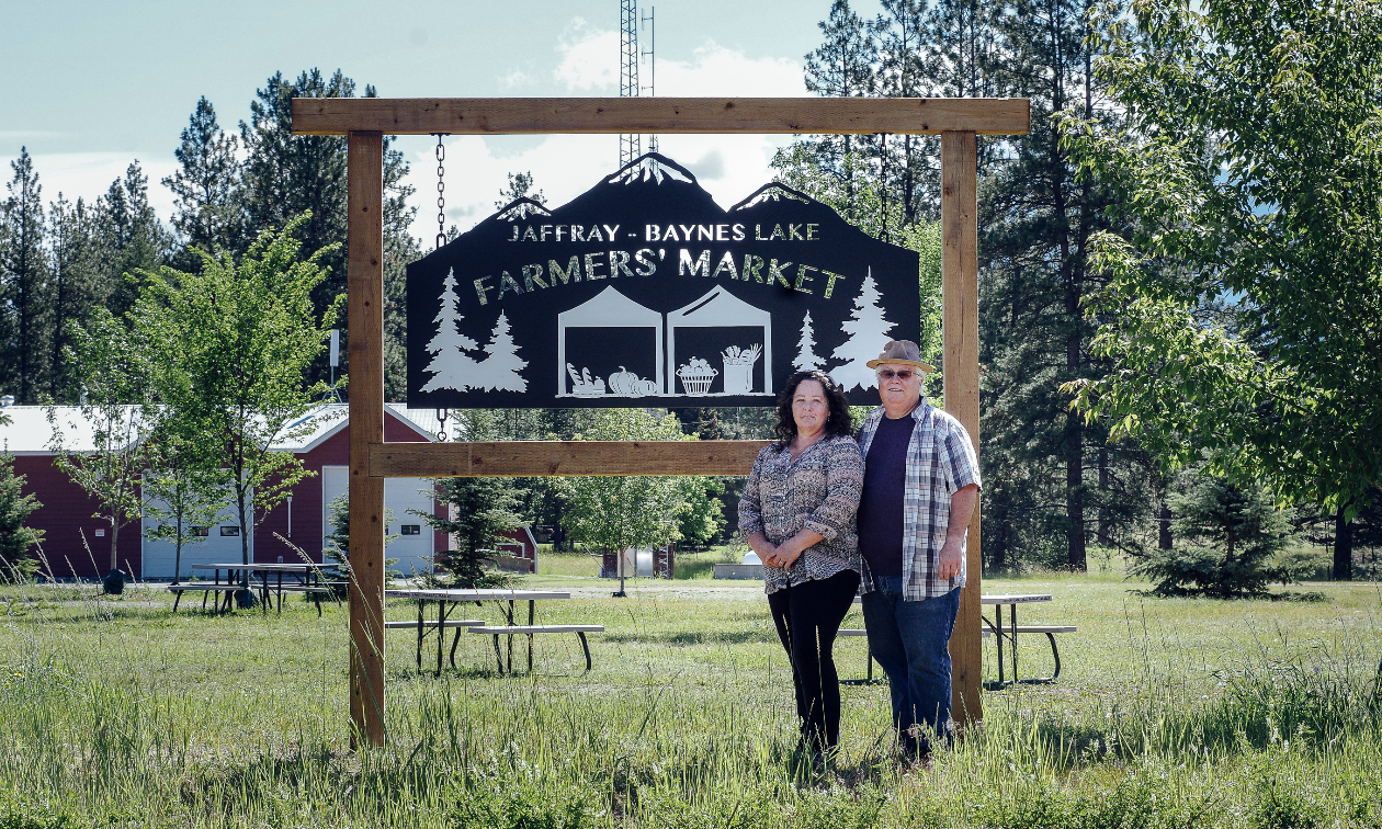 Treanne and Shaun See stand in front of a black and transparent sign that reads Jaffray-Baynes Lake Farmers’ Market. There is greenery in the background as well as a red barn with white doors. 