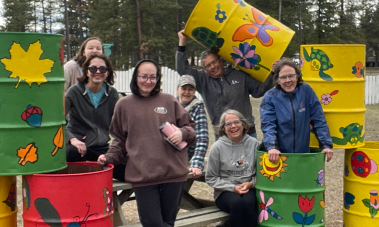 Trash barrels are brightly coloured red, green and yellow with images of leaves and butterflies. Artists stand in and around the barrels with smiling faces. 