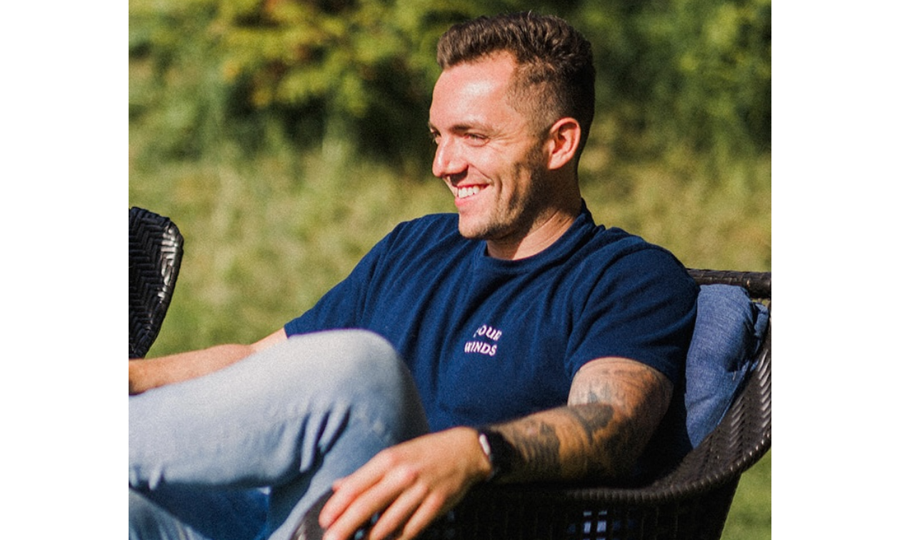 Jeff Kennedy smiles as he relaxes in a wicker chair. He wears light blue jeans and a dark blue T-shirt. He has brown hair and tattoos on his left arm. 