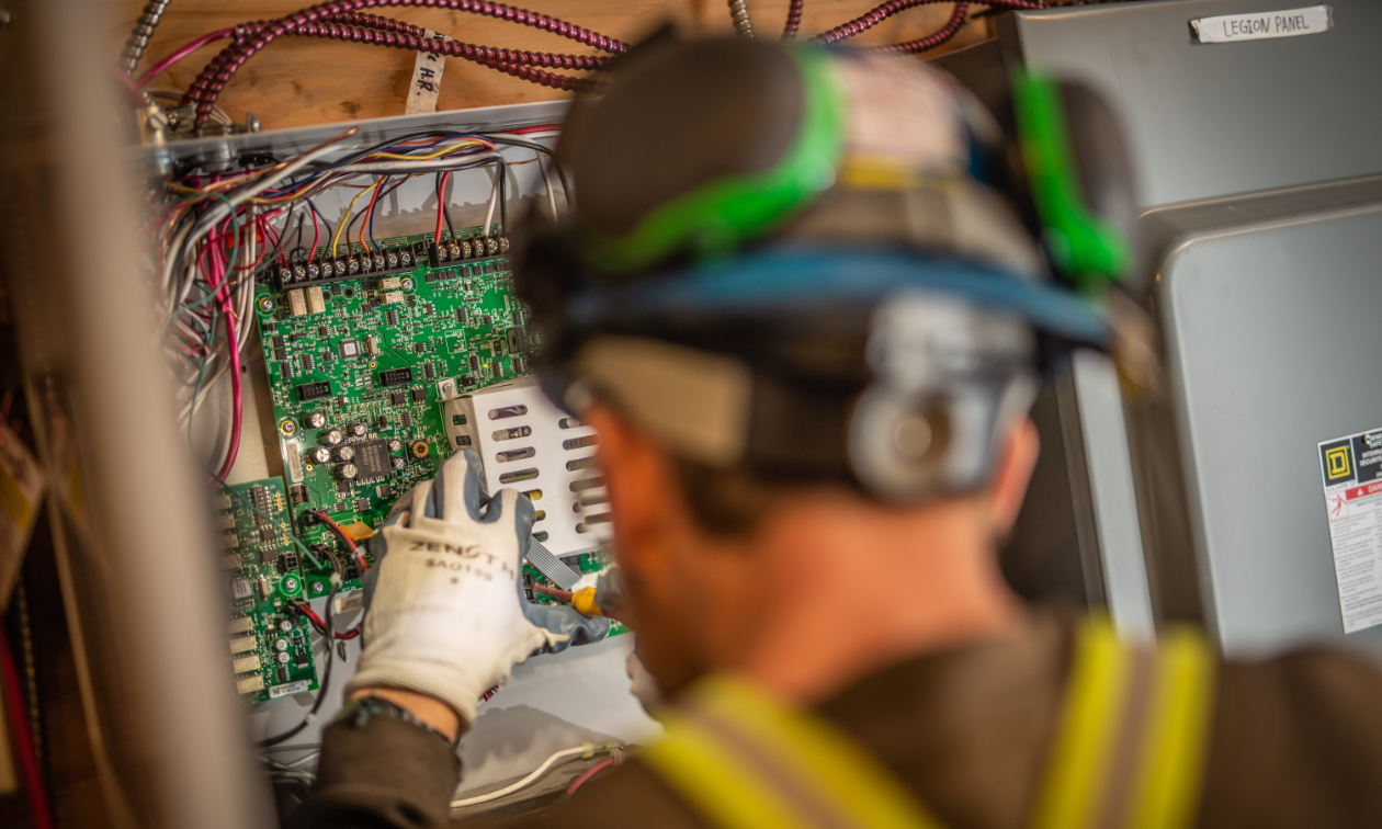 An electrician inspects a circuit board while wearing gloves and a hard hat. 