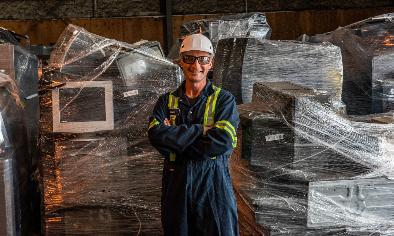 A man smiles wearing a hard hat and protective eyewear in front of a bunch of computers wrapped tightly in plastic wrap.