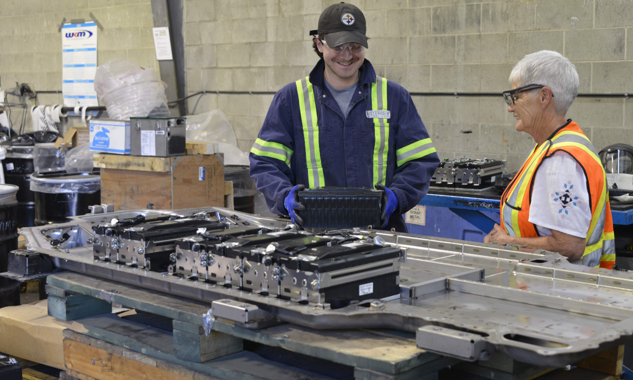 A man and woman smile while working next to each other wearing reflective vests inside of a warehouse.