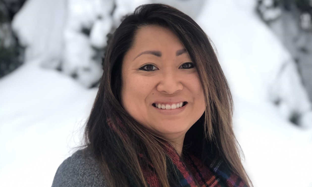 Stacey Brensrud has long brown hair and wears a grey shirt and red scarf. She smiles in the photo. Snowy trees are in the background. 