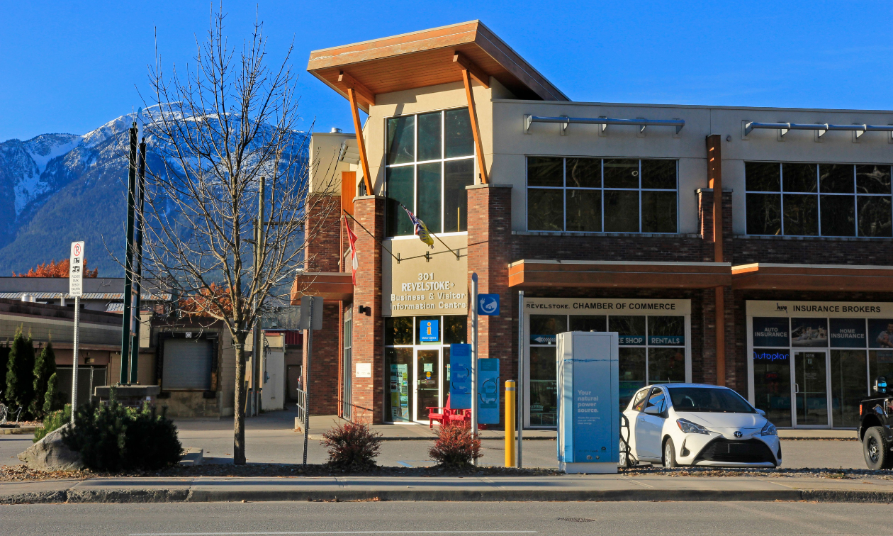 The Revelstoke Chamber of Commerce is a tan and brown building in downtown Revelstoke. Mountains are in the distance. 