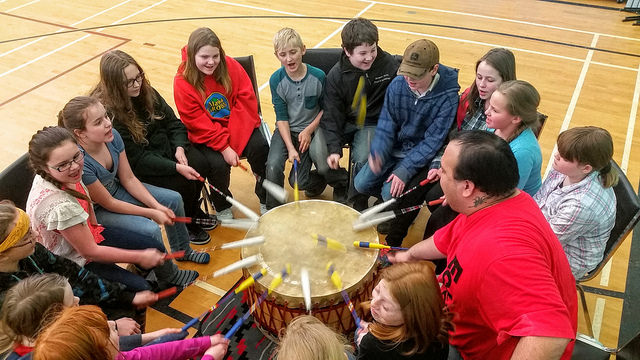 A group of children are sitting in a circle around a drum. All the children have a drumstick in hand and are participating in beating the drum.