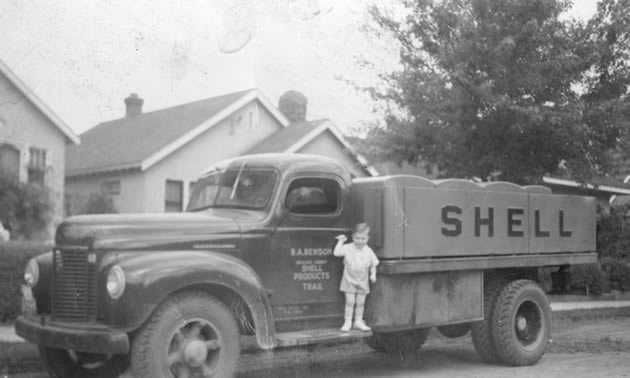 A black and white photo of a young boy standing on the side of an old fuel truck. 
