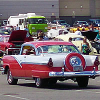 A red and white car surrounded by other classic cars in a parking lot. 