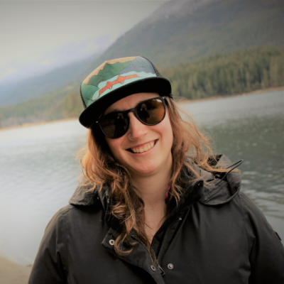 Karen Kornelsen wears a hat and sunglasses with a black coat in front of a blue lake in the woods. 