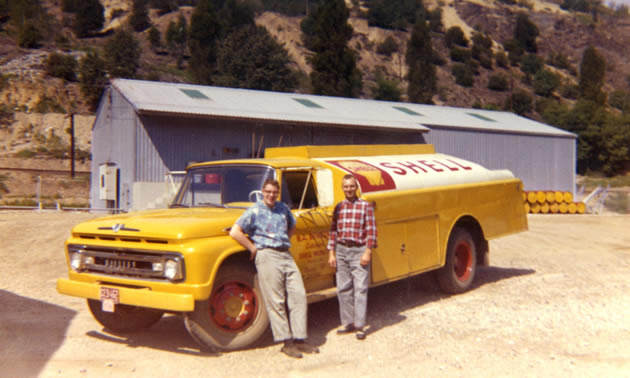 An old photo of two men standing beside a yellow fuel truck. 