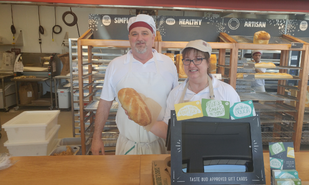 Rod and Tamara Duggan are standing behind the counter of Cobs Bread Bakery and Tamara is holding a loaf of bread.