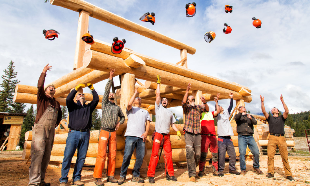 Carpenters throw their hard hats in the air in front of a log building.