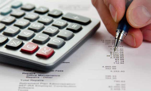 A hand writes on a paper with numbers next to a calculator.