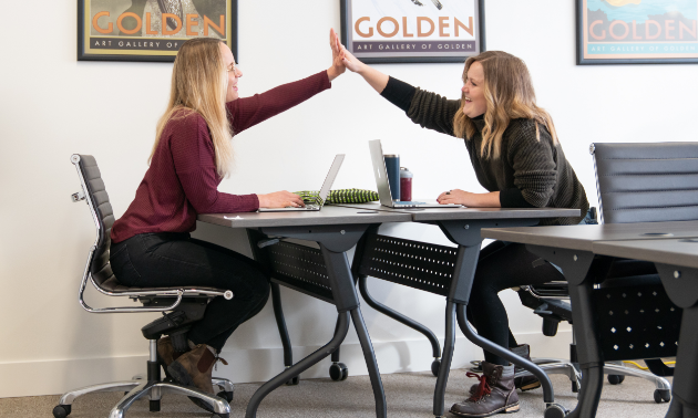 Two women high-five while sitting across from each other at desks.