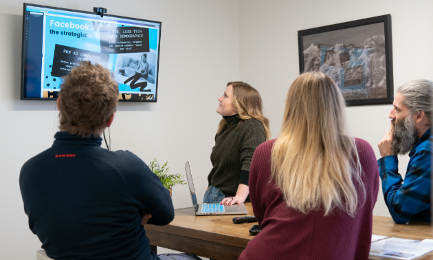 Three people listen to a presentation being presented by a woman at the far end of the room next to a big screen.