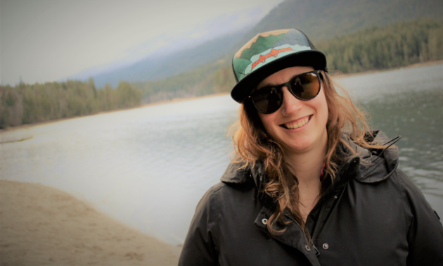 Karen Kornelsen wears a hat and sunglasses with a black coat in front of a blue lake in the woods. 