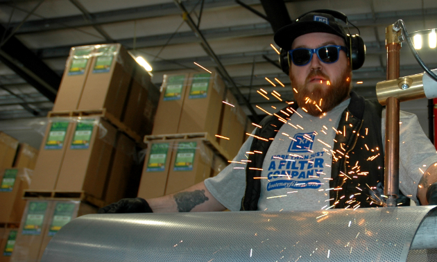 Kyle Grattan wears protective glasses and earmuffs as he works on a machine that sprays sparks.