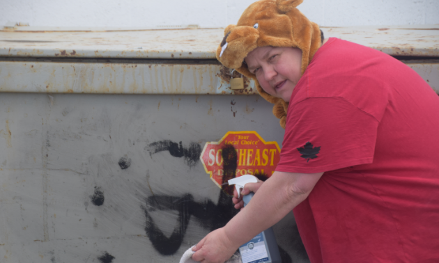 A woman wears a beaver hat costume and red shirt while rubbing off graffiti from a dumpster.