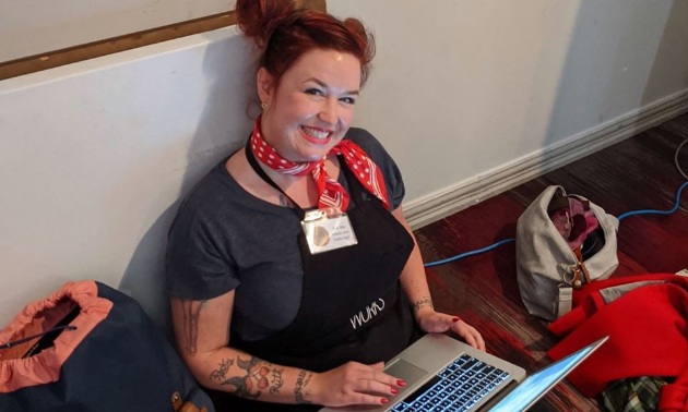 Mandi Lunan sits on the floor wearing an apron while working on her laptop.