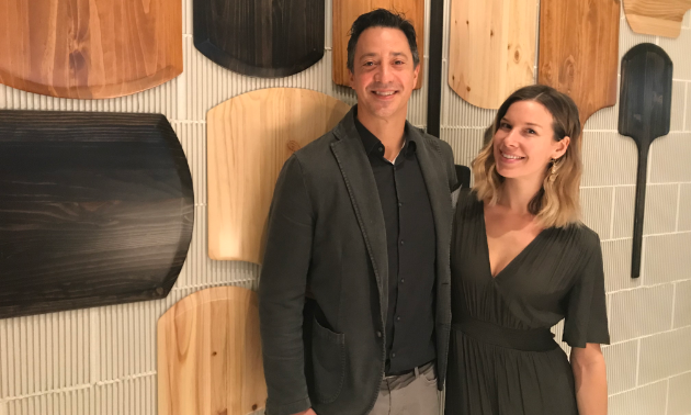 Ryan and Lea Martin, owners of Marzano, stand in front  of a wall with pizza paddles on the wall.