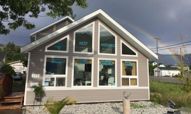 The facade of Rest Assured Kootenay Floats is triangular with many windows on its face. A rainbow climbs over the roof of the house with a darkened sky lingering in the distance.