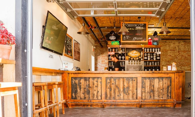 The taproom of Rossland Beer Company has a wooden bar with tan, wooden stools set to the side. A big widescreen TV hangs on the wall. Upside down cone lamps hang from the ceiling.