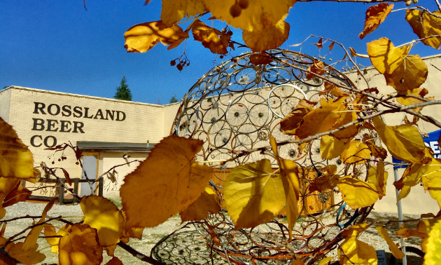 The front of Rossland Beer Company says the business’ name in dark lettering on a beige building. In the foreground, yellow fall leaves are in front of a transparent, decorative golden sphere.