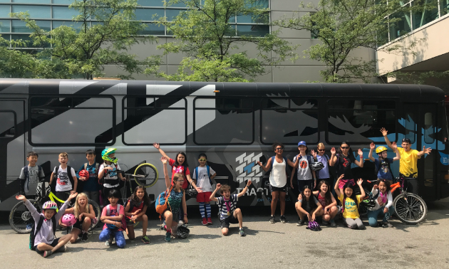 Kids line up in front of a big black bus and wave to the camera.