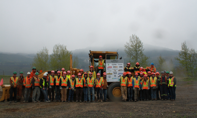 Students wearing orange and yellow vests pose in front of heavy machinery for Worksafe BC