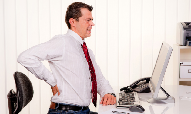 A man grabs at his back in pain while sitting at a desk