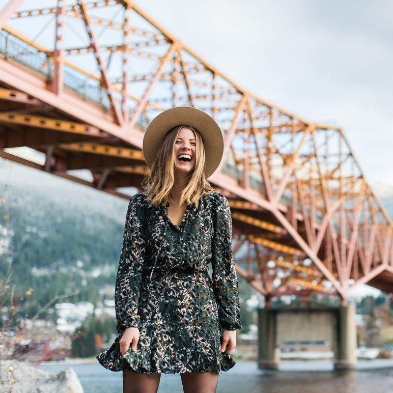 Amanda Mary laughs while wearing a dress in front of an orange bridge in Nelson, B.C.