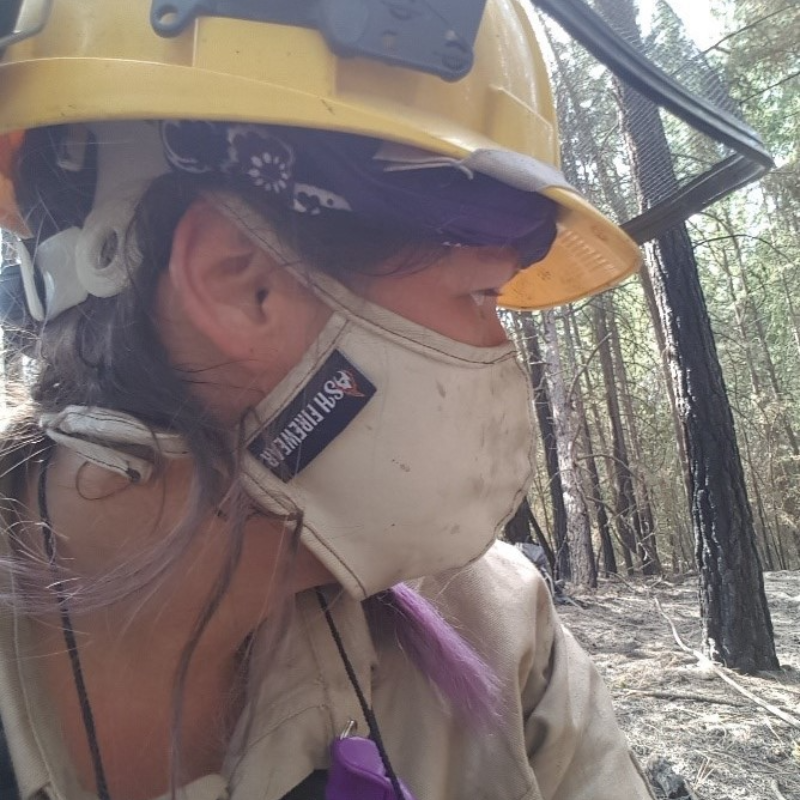 A female firefighter wears a yellow hard hat and a mask that says AshFireWear.