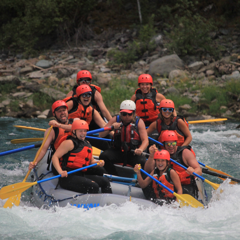 A group of white water rafters smile as they dip into deep water on a raft. 