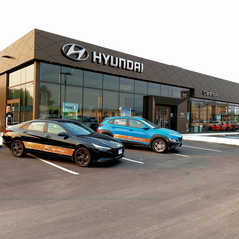 Two cars, one black, one blue, are parked in front of Cranbrook Hyundai’s new showroom with large glass windows. 