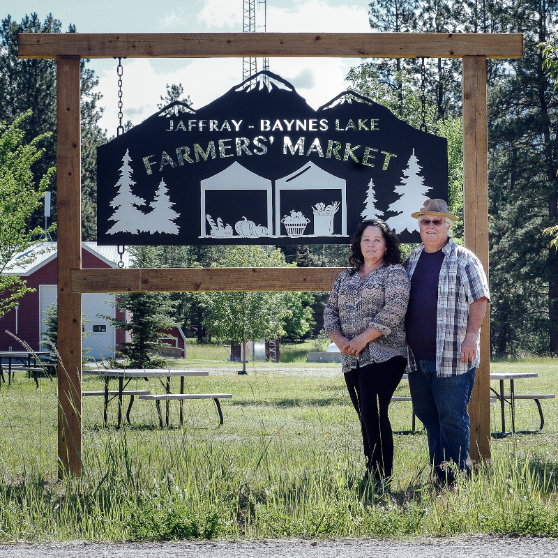 Treanne and Shaun See stand in front of a black and transparent sign that reads Jaffray-Baynes Lake Farmers’ Market. There is greenery in the background as well as a red barn with white doors. 