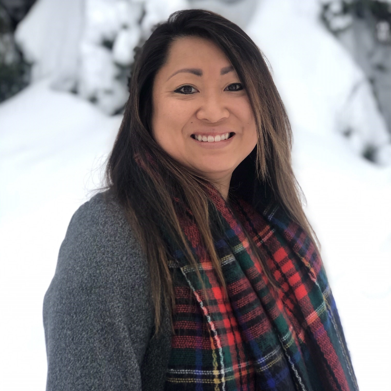 Stacey Brensrud has long brown hair and wears a grey shirt and red scarf. She smiles in the photo. Snowy trees are in the background. 