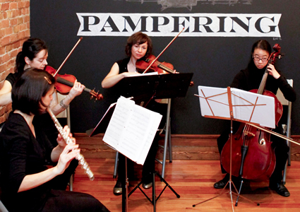 Four young girls playing different instruments in front of a wall with a banner on a hardwood floor stage.