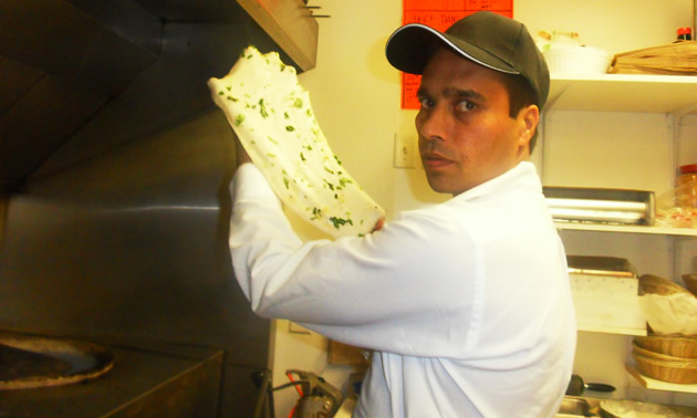 Man wearing a baseball cap and chef's whites with a flatbread-type sheet of dough in his hands 
