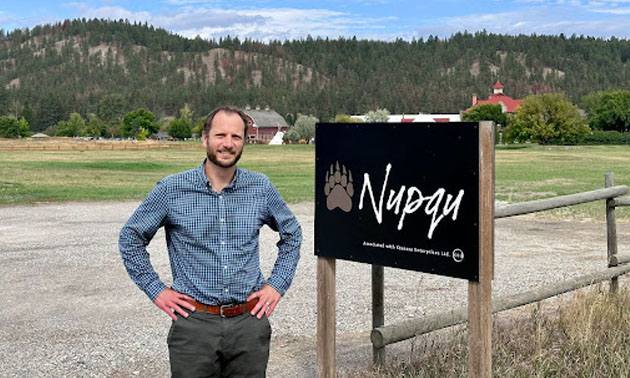 Aaron Higgs, standing beside Nupqu Resources sign. Treed mountain in background. 