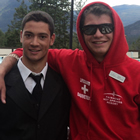 Two smiling young men, one in a black suit and one in a red hoodie, pose for the camera 