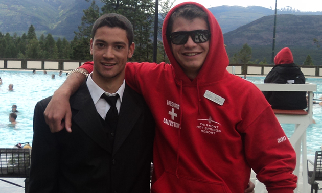 Two smiling young men, one in a black suit and one in a red hoodie, pose for the camera 