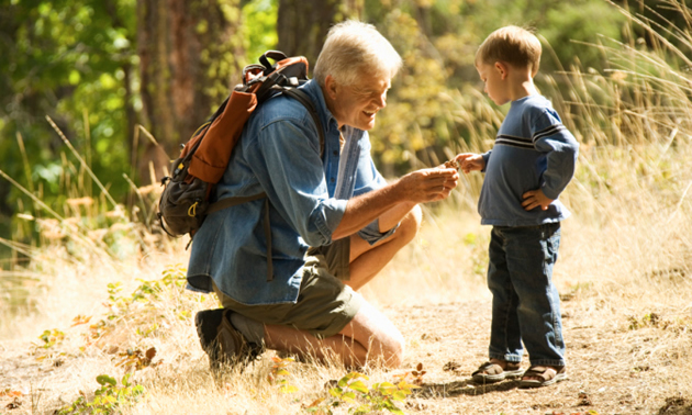 A grandfather with a backpack on kneels down to show a boy something on his hand. They are on a hike.